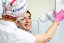 Female Dentist Is Going To Make Teeth X-ray Image For Handsome Young Man In Dental Clinic. Using Dental X-ray Machine In Dental Office. Dental Equipment Stock Photo, Picture And Royalty Free Image.